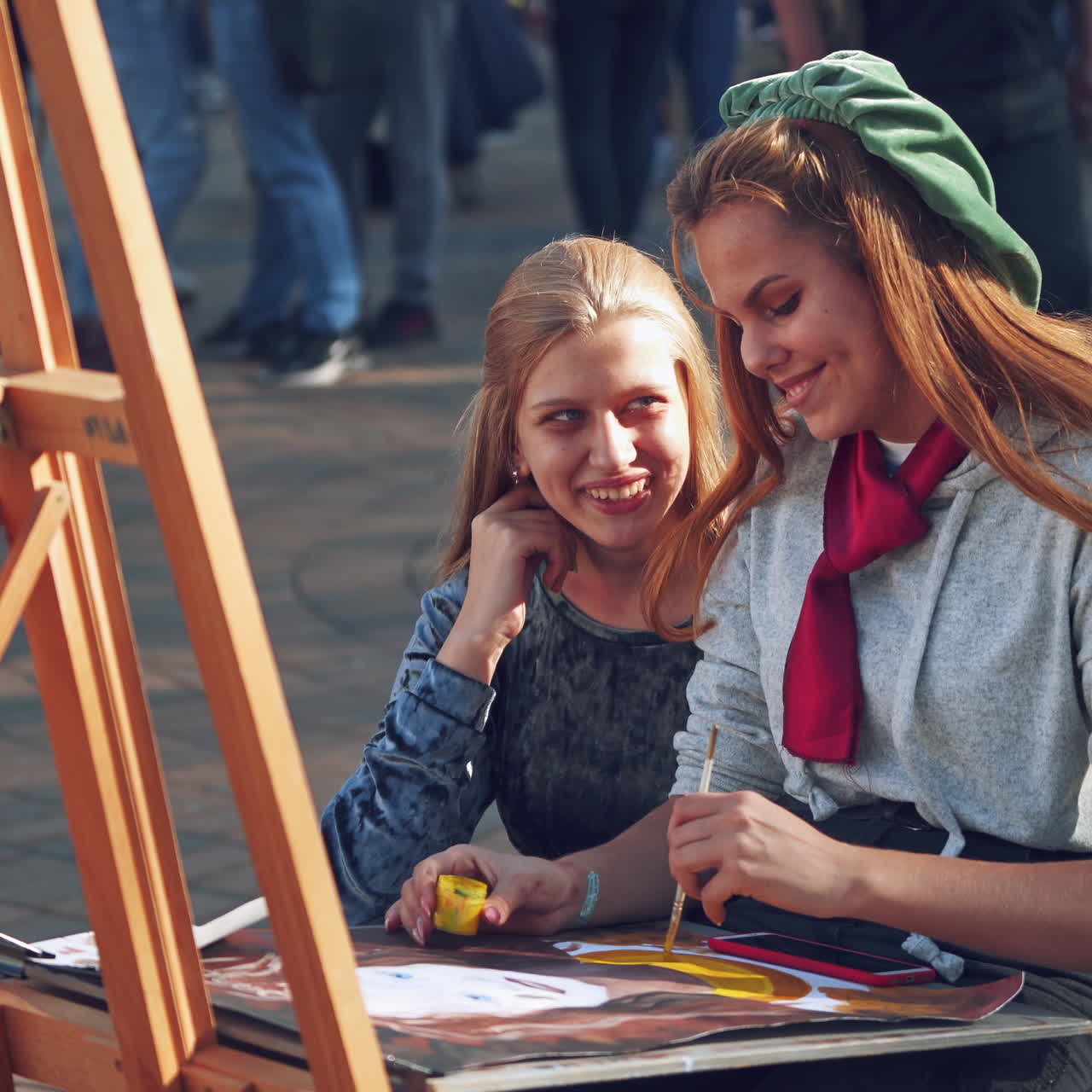 Two women near easel. Beautiful girl artist painting with brush on the busy street background. Female friend talking to female artist outdoors.