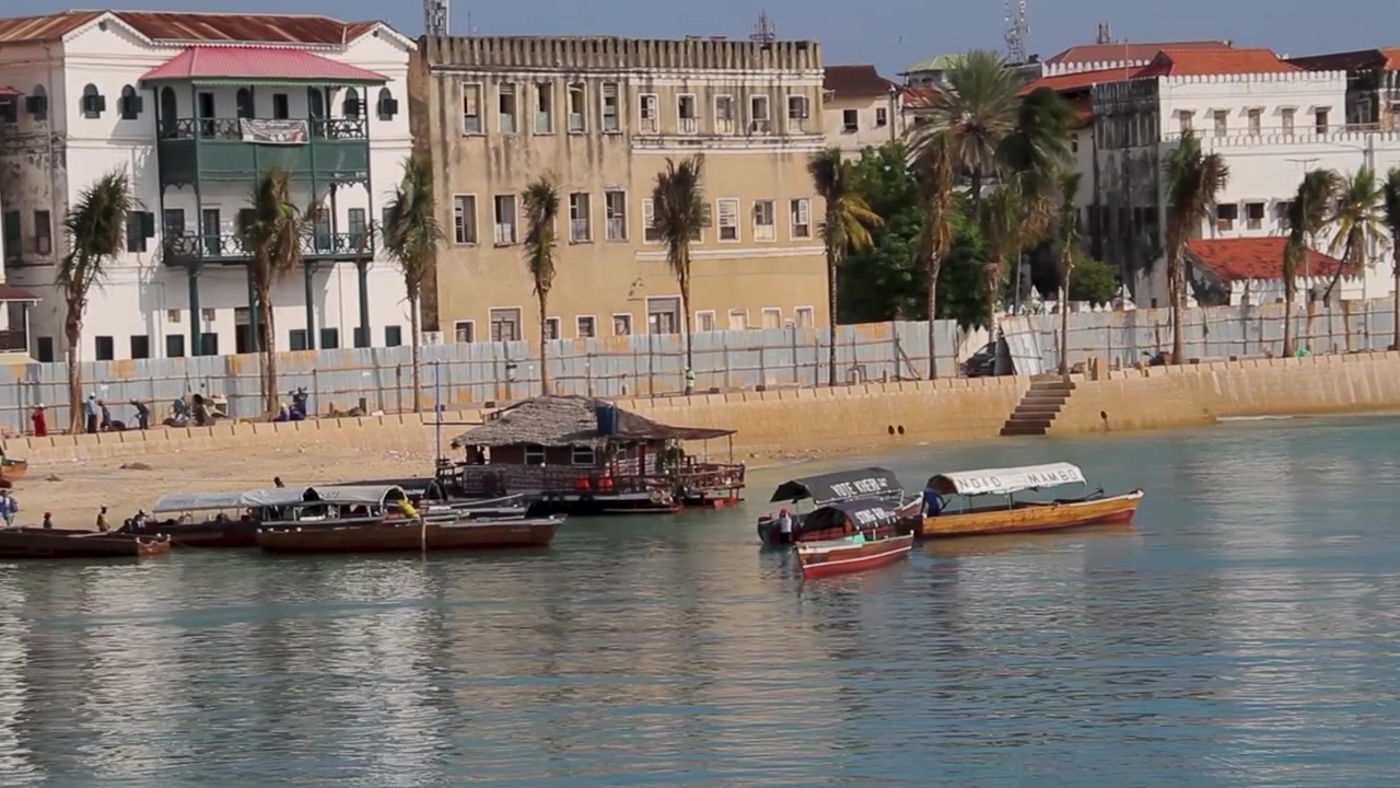 Pemandangan indah perahu dhow kayu tradisional yang berlabuh di sepanjang pantai berpasir Stone Town, Zanzibar, dengan arsitektur kolonial bersejarah di latar belakang