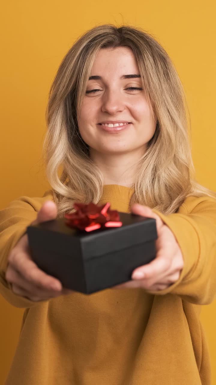 Happy young woman receiving gift box and saying thanks on yellow background