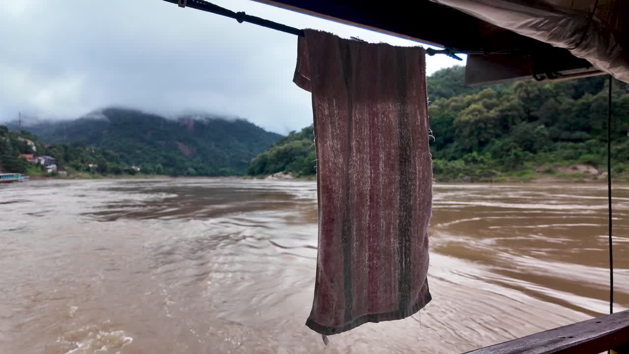 En handduk hänger på aktern av en långsam båt som färdas på Mekongfloden i Laos med ett bergigt landskap och molnig himmel i bakgrunden