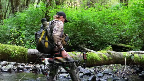 male hunter with a bow crosses a small river creek