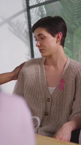 Vertical-Video-Of-Young-Woman-Wearing-Pink-Breast-Cancer-Awareness-Ribbon-Being-Comforted-At-Meeting-Of-Therapy-Support-Group-For-Cancer-Treatment-Patients-With-Counsellor