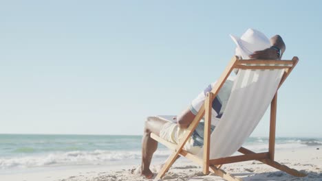 happy senior african american man sitting on deck chair at beach, copy space, in slow motion