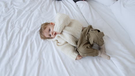 little caucasian toddler boy wearing white knitted sweater and sport pants lies on the bed. lovely kid is resting holding a finger at his mouth. parents sit beside at the big bed. top view.