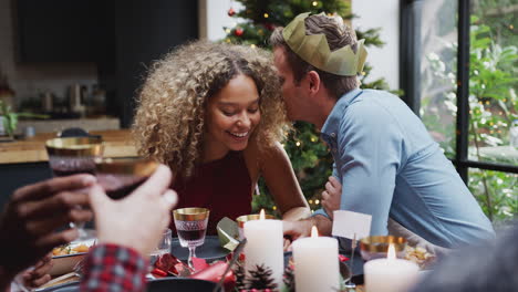 Couple-Sitting-Around-Dining-Table-At-Home-For-Christmas-Dinner-With-Friends-Talking-And-Whispering