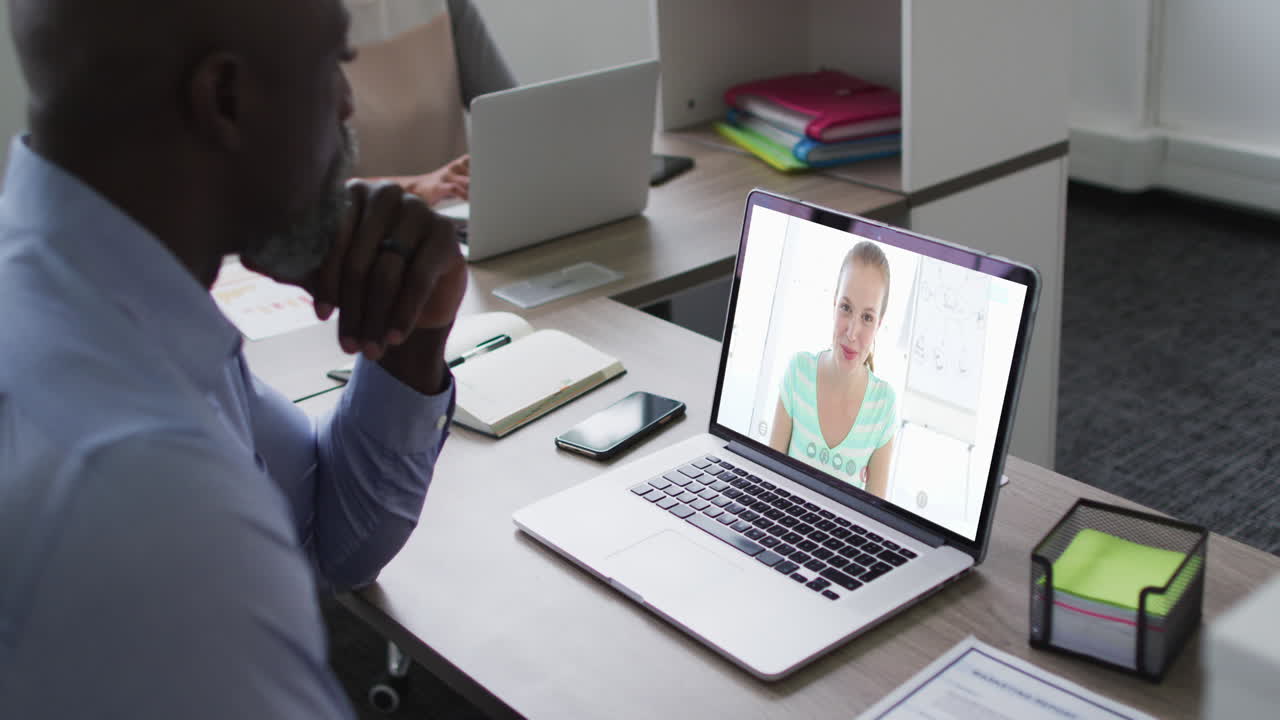 Afroamerikanischer älterer Mann bei einem Videoanruf am Laptop mit einer weiblichen Bürokollegin im Büro