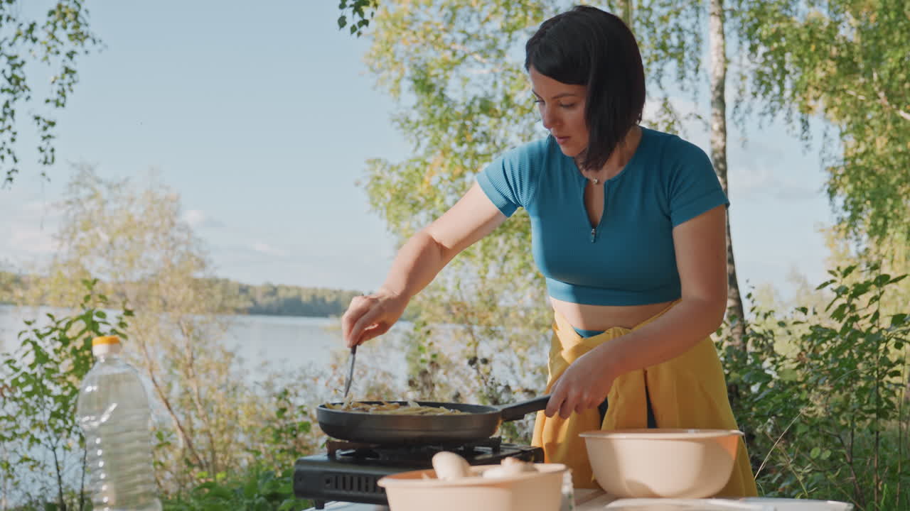 Woman stirring skillet by lake, summer breeze. Sunlit shoreline with birch trees and wide water view, portable gas stove on table, spatula mixing ingredients, fresh produce and bowls, upbeat outdoor