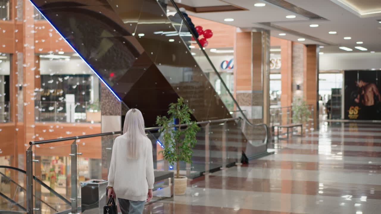 elegant shopper strolls along mall towards bench by moving walkway, opens bag to retrieve book, reads intently, flips through pages as fellow patrons descend escalator in retail atrium setting