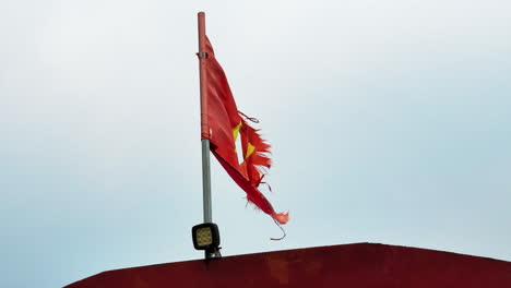 frayed red vietnamese flag with faded yellow star flaps in the wind above a rusted barge roof under a pale sky in the hòa bình reservoir area, northern vietnam, recorded in real time.