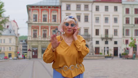 senior old tourist woman in stylish clothes talking on mobile phone while walking on city street