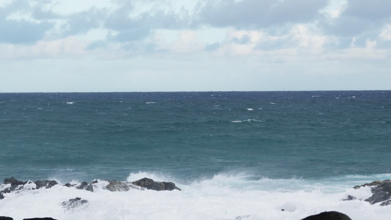 Ocean waves surge and break against jagged black rocks, sending white foamy spray into the air along a rugged shoreline under a cloudy horizon