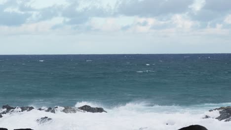 ocean waves surge and break against jagged black rocks, sending white foamy spray into the air along a rugged shoreline under a cloudy horizon