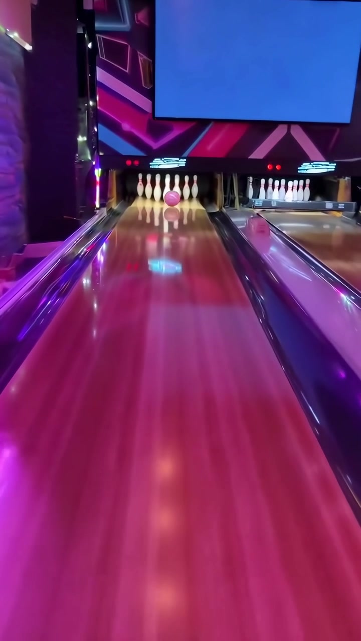 Růžová bowlingová koule kutálející se po neonově osvětlené dráze a srážející kuželky.