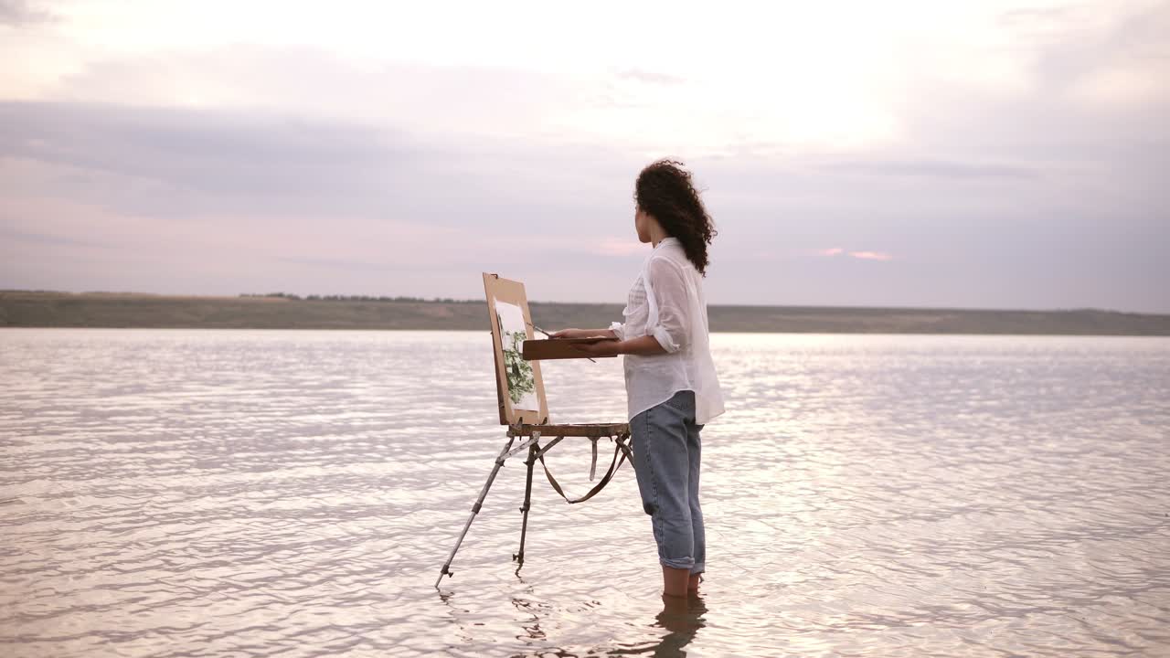 Accelerated shooting, a girl artist in the water till ankles draws a landscape using an easel and a palette. Curly girl on the background is lake, she's in casual clothes: jeans and a white shirt