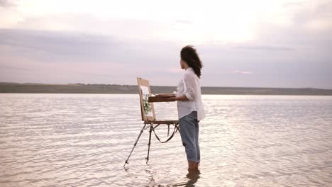 accelerated shooting, a girl artist in the water till ankles draws a landscape using an easel and a palette. curly girl on the background is lake, she's in casual clothes: jeans and a white shirt