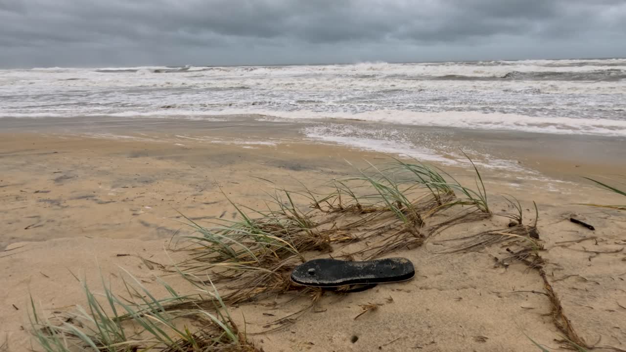 흐린 하늘 아래 잔해와 강한 파도가 있는 폭풍우가 몰아치는 해변 장면, 환경 영향과 해안 침식에 중점을 둔다.