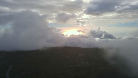 Drone-footage-of-a-moutain-in-the-clouds-with-a-small-road-and-a-sunrise