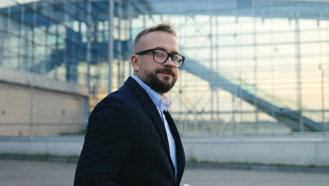 Side-View-On-The-Handsome-Businessman-In-Glasses-Walking-Outside-Big-Glass-Urban-Building-Of-Airport,-Drinking-Coffee-And-Turning-His-Head-To-The-Camera
