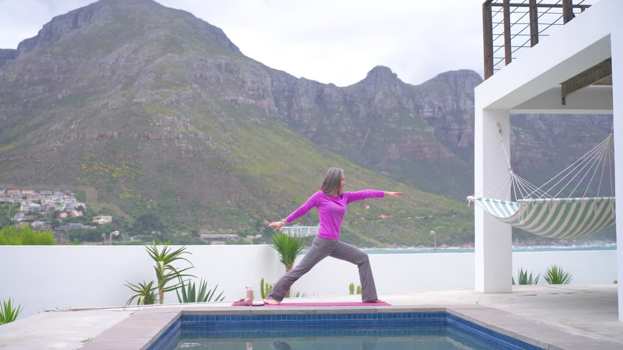 Mature woman flowing from lunge into warrior sequence on pink mat at pool terrace for balance