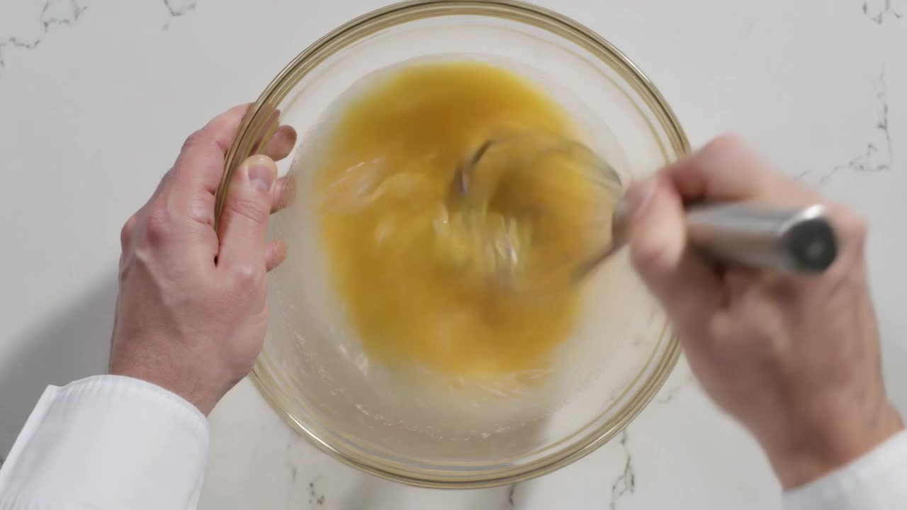 Chef con polsino bianco che inserisce una frusta in una ciotola, sbattendo un composto giallo su un bancone di marmo per amalgamare.