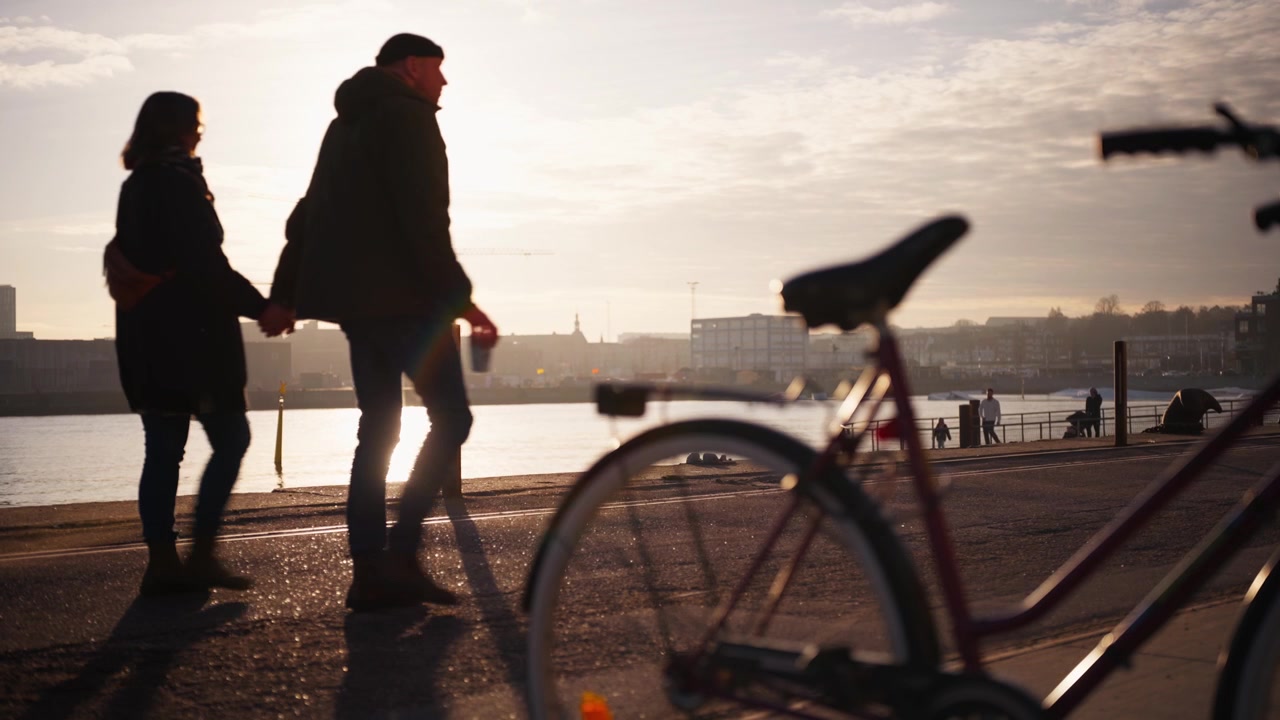 Nærbilde av en sykkel i silhuett mot en gyllen solnedgang på en strandpromenade, med kraner og fotgjengere i det fjerne