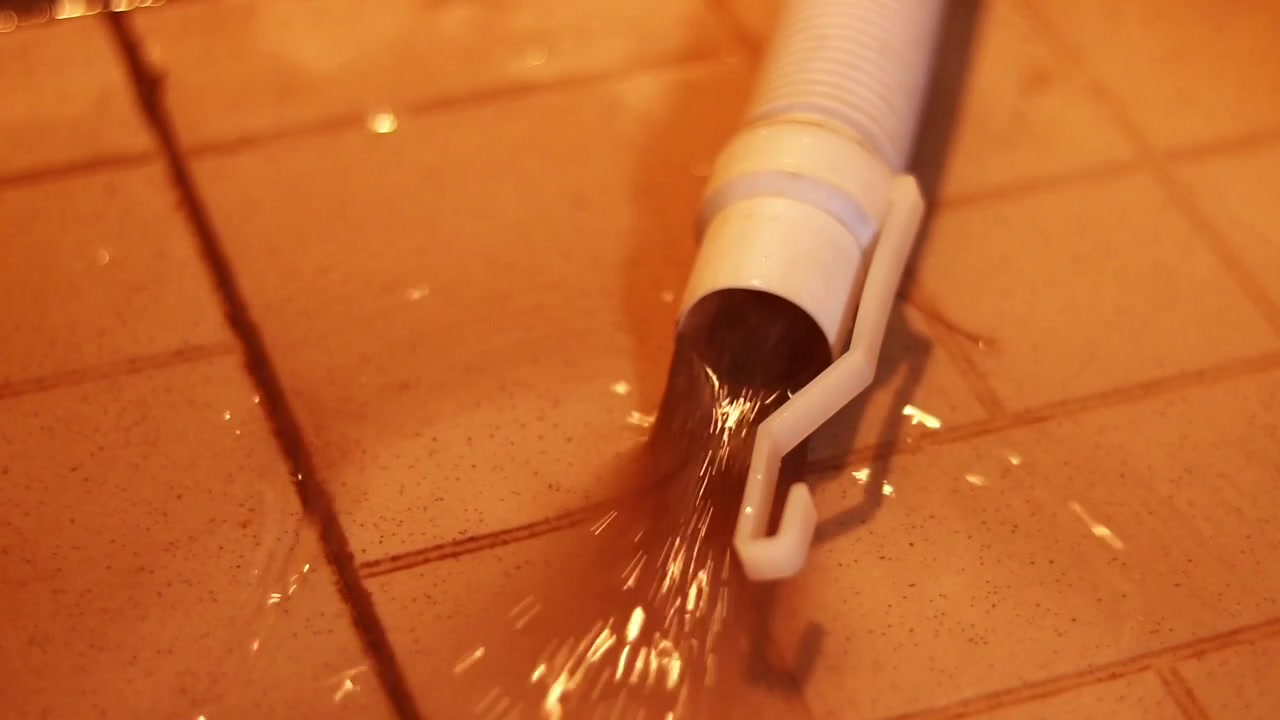 Flexible white drain hose empties a steady stream of water across glossy brown tiles under warm indoor lighting, with ripples and reflections spreading outward.