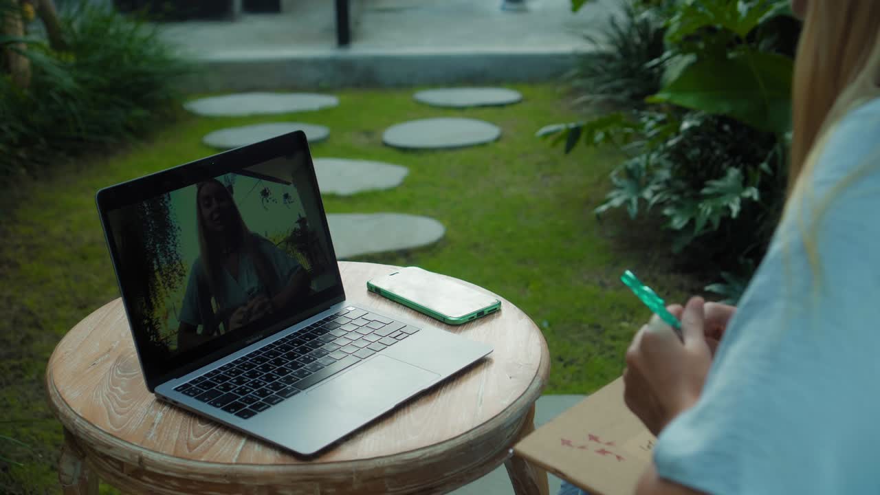 una chica en un vestido azul remotamente en línea trabajando detrás de una computadora portátil y mirando a la pantalla el patio trasero con plantas verdes