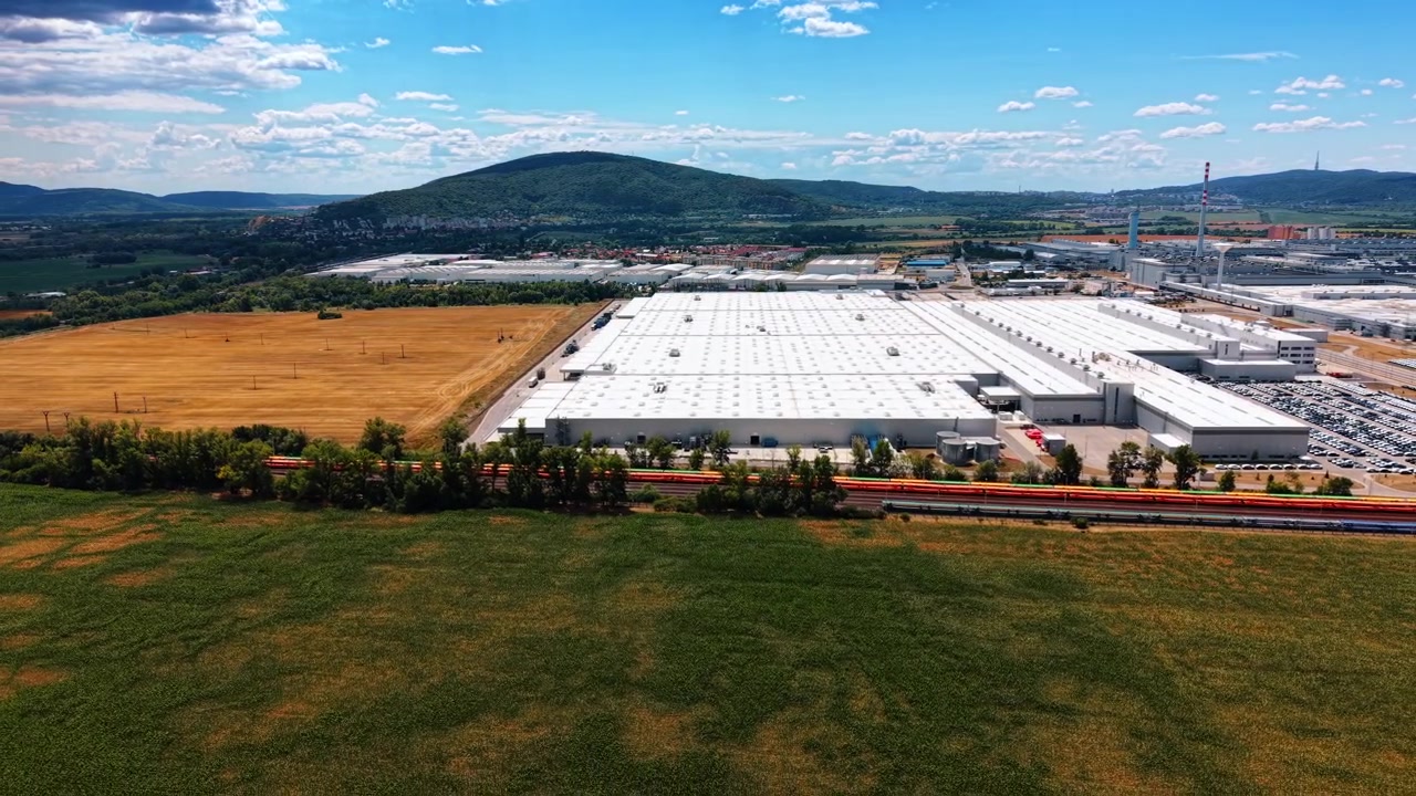 Magazzino industriale tra campi agricoli e ferrovia. Vagoni colorati passano una fila di alberi sotto ampi tetti bianchi, serbatoi di stoccaggio e colline boscose in lontananza