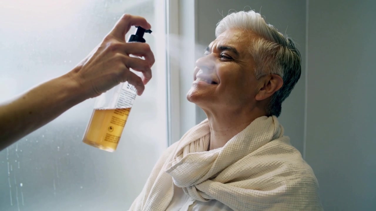 Spraying stylist pressing nozzle, man with towel tilting into mist near frosted glass for grooming