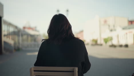 rear view of a woman sitting outdoors on a wooden chair in urban setting, wearing black coat, with blurred pedestrians walking by and modern city architecture in soft daylight