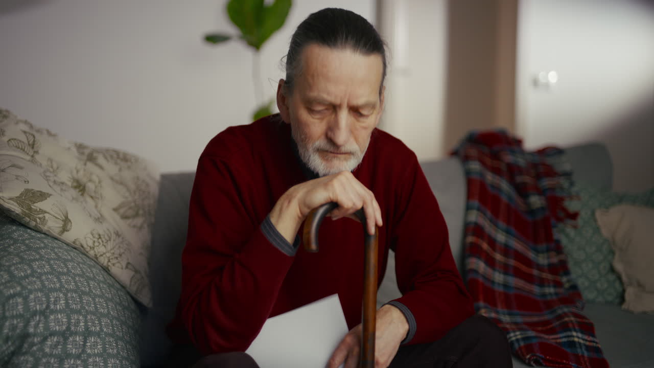 Sad Elderly Man Sitting on Couch with Walking Stick and Paper