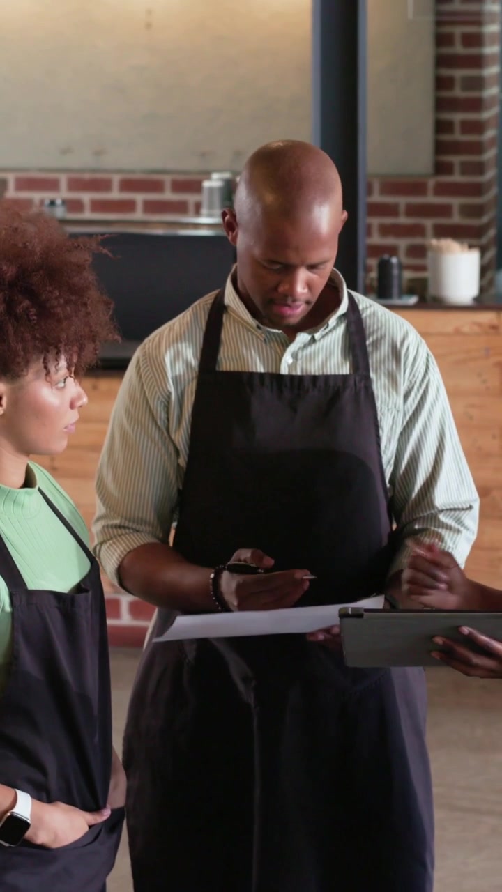 Vertical video: Reviewing two staff studying clipboard and tablet pre-shift at cafe wearing aprons