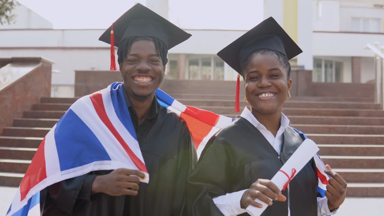 hombre y mujer afroamericanos de pie uno al lado del otro frente a la cámara con túnicas negras y sombreros cuadrados de estudiantes graduados con la bandera británica en los hombros