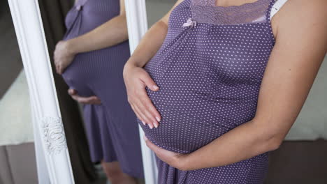 beautiful pregnant lady in a nightdress standing in front of a mirror and looking at her abdomen. future mom caresses her future child in the tummy. close up.