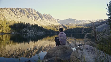 person sitting by a tranquil mountain lake, surrounded by nature in golden sunlight