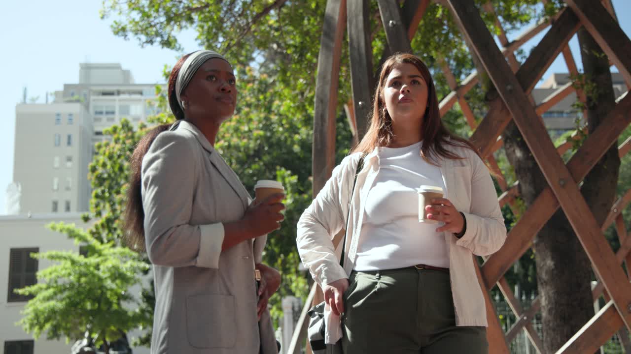 Diverse donne conversano sotto un pergolato di legno in una piazza; una a destra solleva una tazza leggendo il cellulare, mentre un'altra a sinistra controlla l'orologio.
