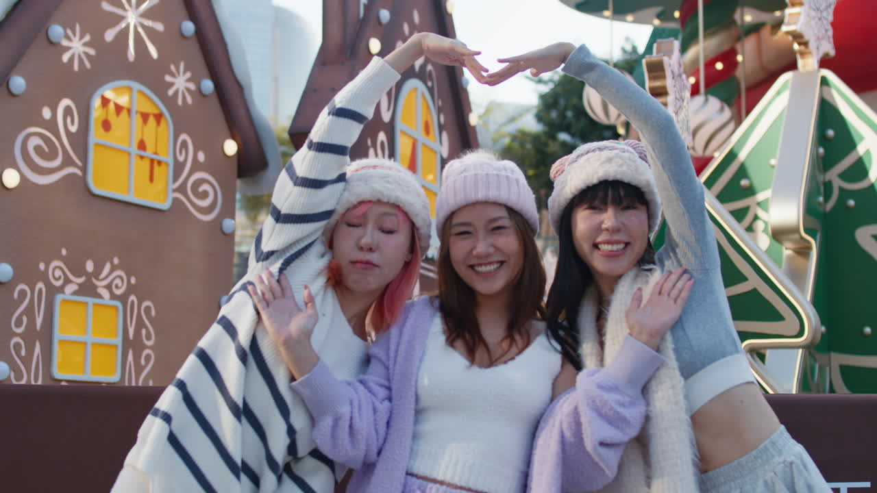 Three smiling women posing in front of a gingerbread house during the winter