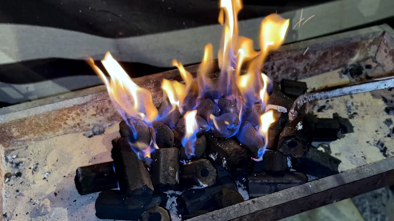 Close-up of metal tongs arranging glowing hexagonal charcoal briquettes in a sand-lined grill. Blue and orange flames rise from the hot coals, creating intense heat for outdoor cooking or BBQ