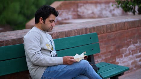 Ratón-De-Biblioteca-Masculino-Disfrutando-De-La-Naturaleza-Mientras-Lee,-Joven-Del-Sur-De-Asia-Absorto-En-La-Lectura-Al-Aire-Libre