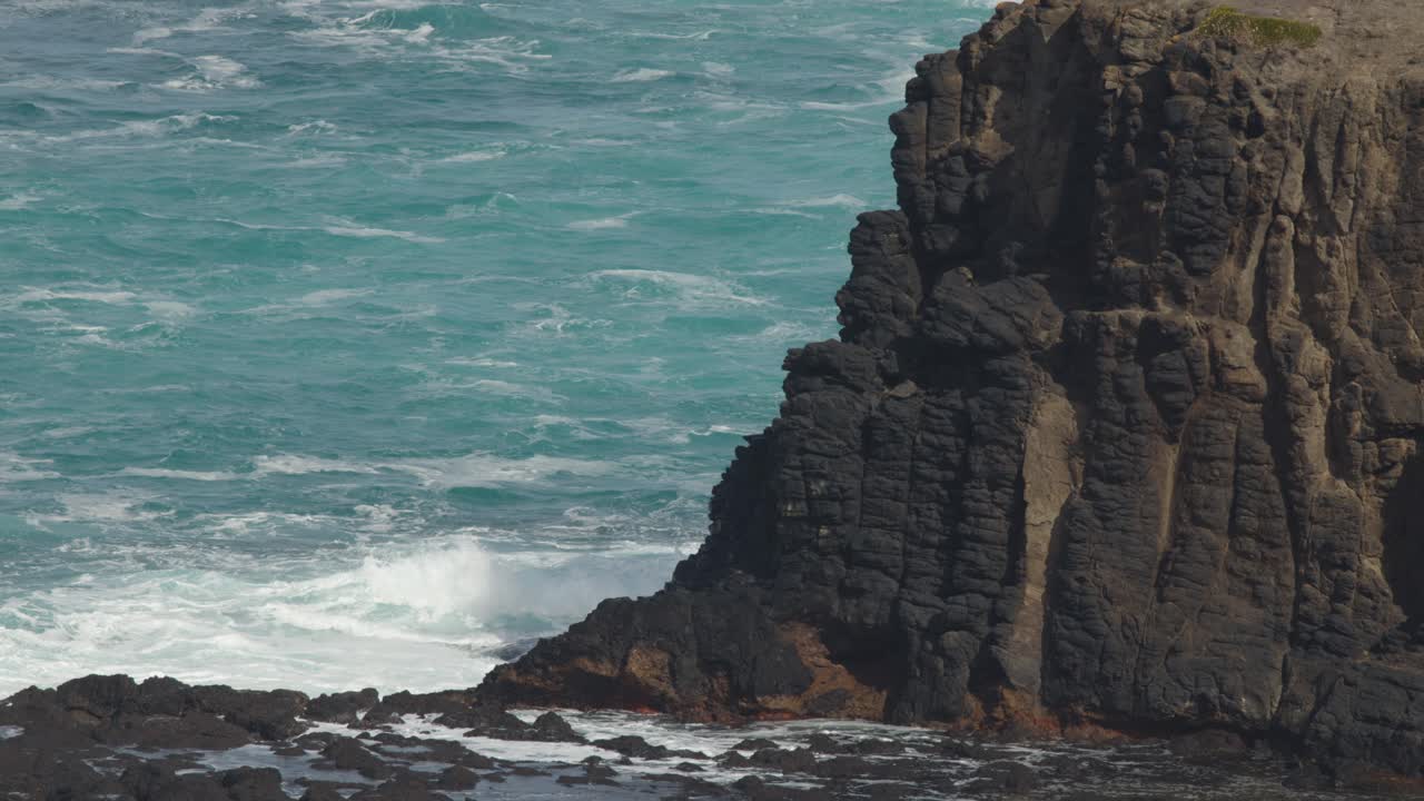 Ocean waves hit rugged basalt cliffs under daylight, static camera, natural coastal landscape, dramatic mood