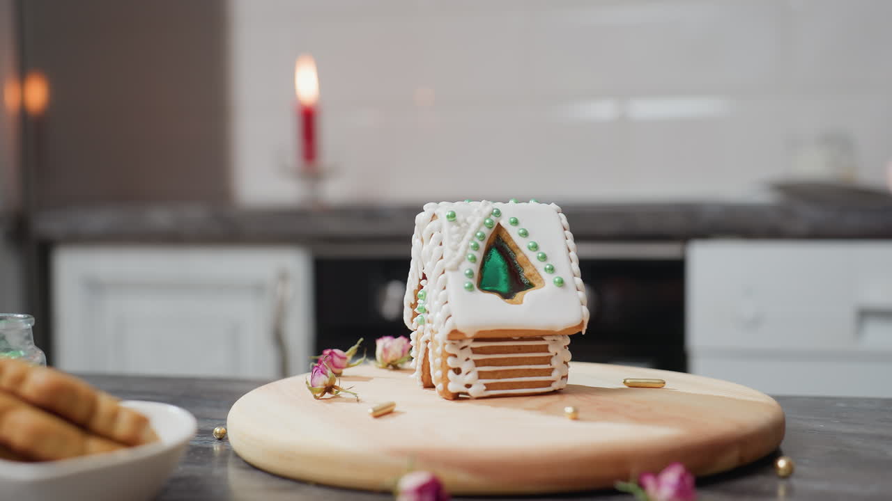 Hand carefully placing green bead on gingerbread house decorated with white icing, wooden tray, dried flowers, golden sprinkles, and blurred candlelight in background