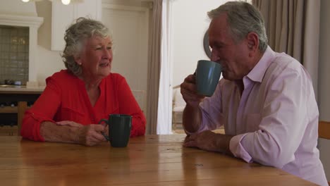 una pareja de ancianos caucásicos sonrientes sentados a la mesa en el comedor, hablando y bebiendo tazas de café