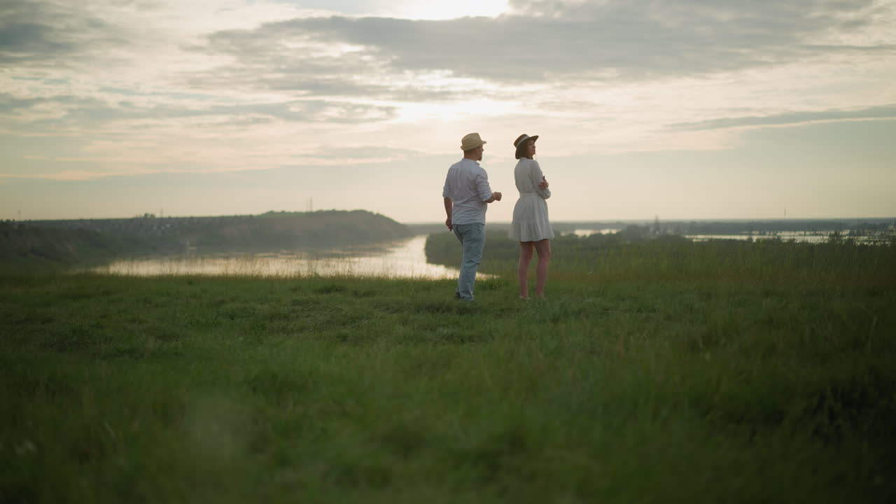 모자,  셔츠, 청바지를 입은 남자가  드레스를 입은 여자를 향해 걸어 가며, 해가 지는 동안 호수를 내려다보는 잔디 언덕에 서 있습니다.