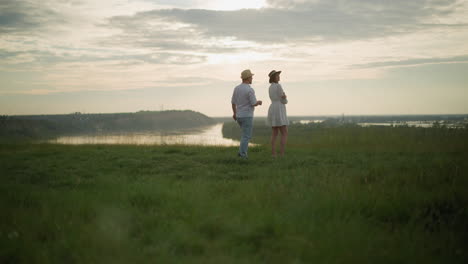 un hombre con sombrero, camisa blanca y vaqueros camina hacia una mujer con un vestido blanco de pie en una colina cubierta de hierba con vistas a un lago al atardecer. momento con un hermoso paisaje