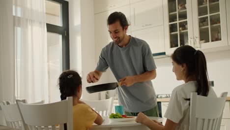 Ein-Brünetter-Mann-In-Einem-Grauen-T-Shirt-Legt-Seiner-Frau-Und-Seiner-Kleinen-Tochter-Beim-Frühstück-In-Der-Küche-Am-Familientisch-Morgens-Aufgewärmten-Toast-Aus-Der-Pfanne