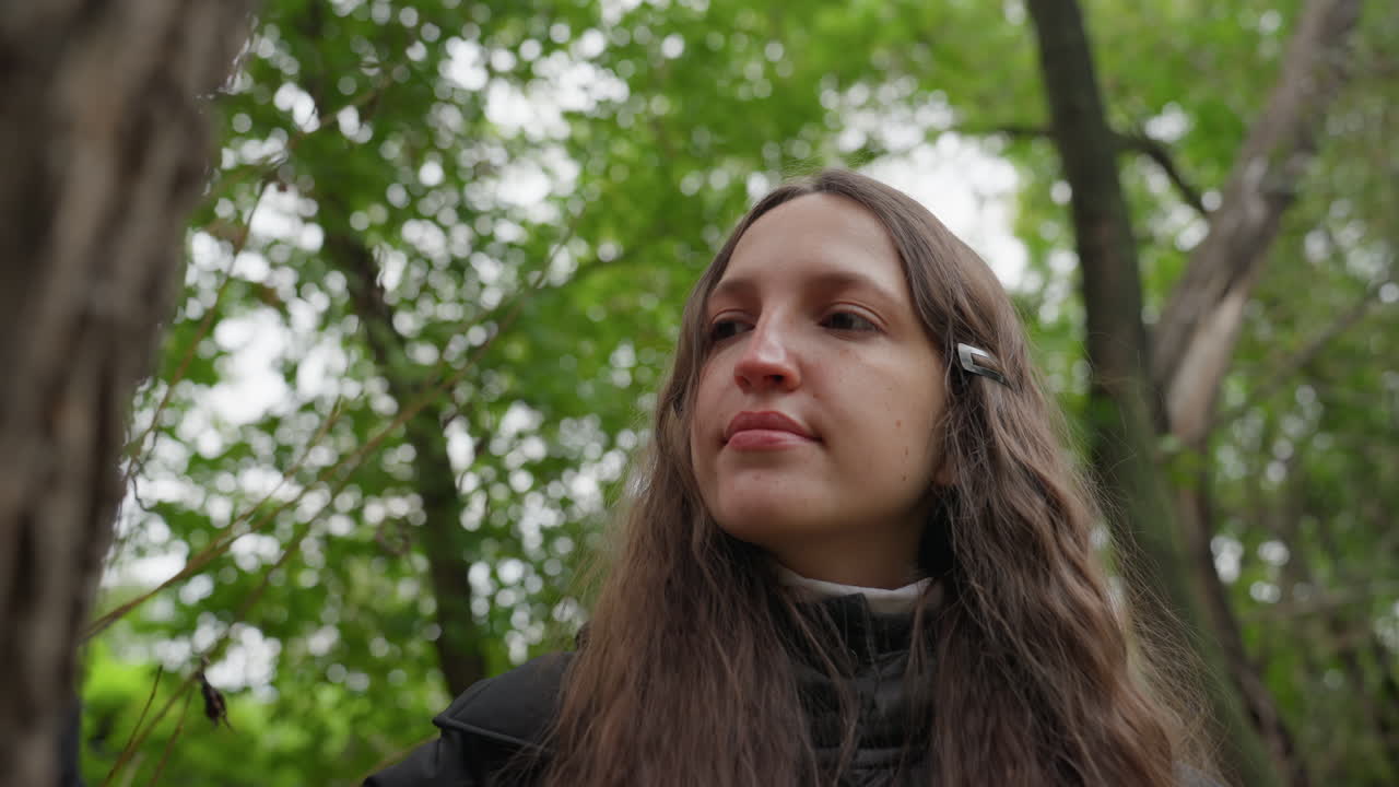 Una donna con una giacca nera si gode la natura. Una giovane donna tranquilla percorre con calma un sentiero nel bosco. Scena pacifica di una giovane donna con lunghi capelli scuri nella foresta, circondata dalla luce solare screziata.