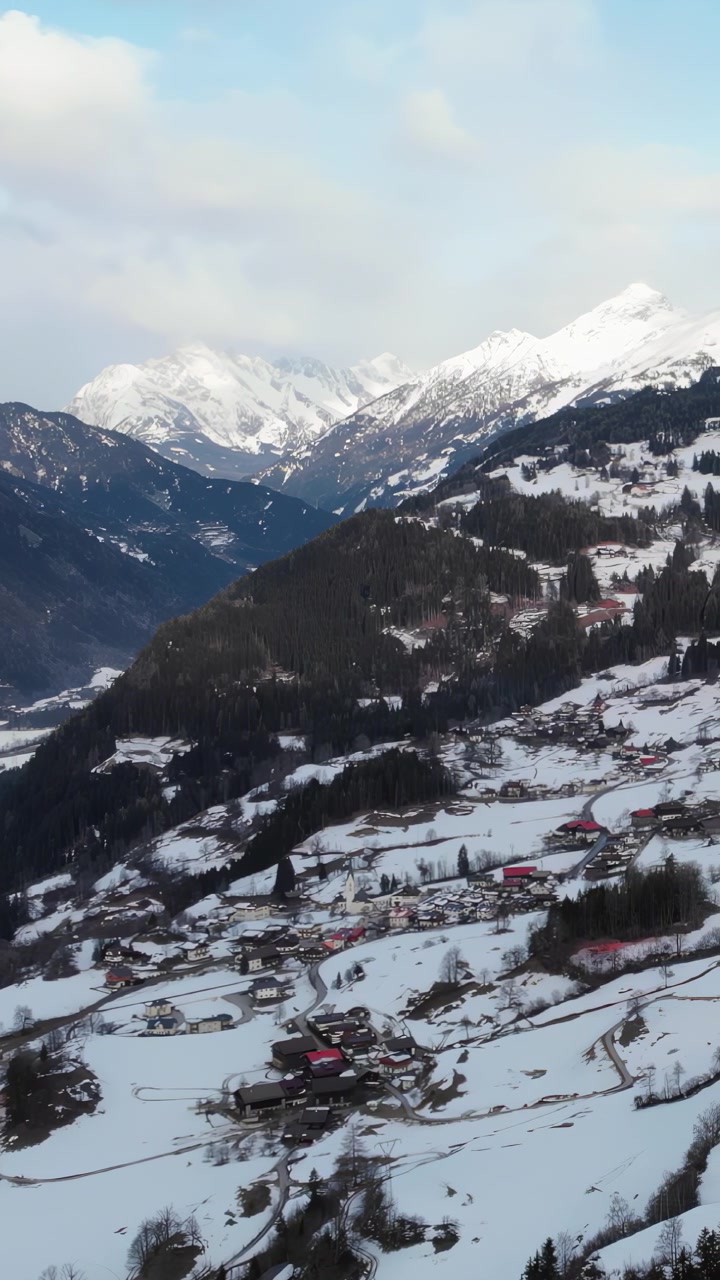Ośnieżona alpejska wioska położona w dolinie, dron przelatuje nad polami tarasowymi i krętą drogą, rozproszone chaty