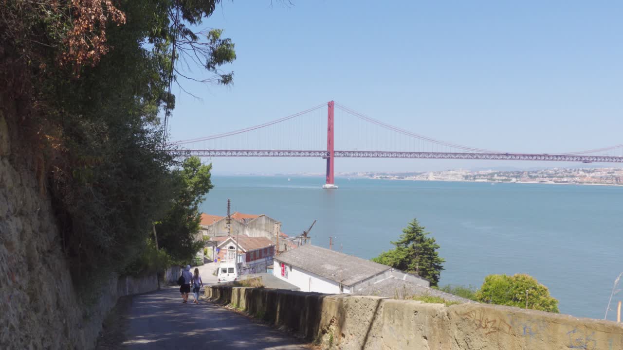 Una coppia che percorre la strada verso un villaggio di pescatori sull'altra sponda del fiume a Lisbona, con il Ponte 25 de Abril visibile sullo sfondo