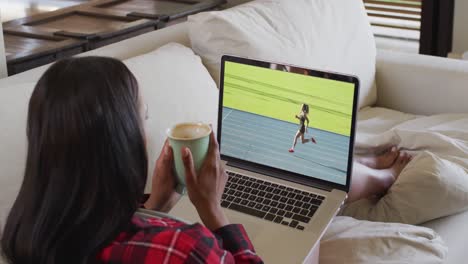 Composite-of-woman-sitting-at-home-holding-coffee-watching-athletics-running-event-on-laptop