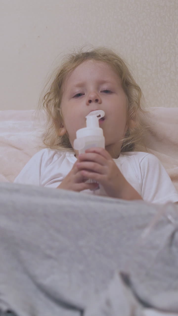 worried ill little girl with modern inhaler lies in comfortable bed near beige wall in light room